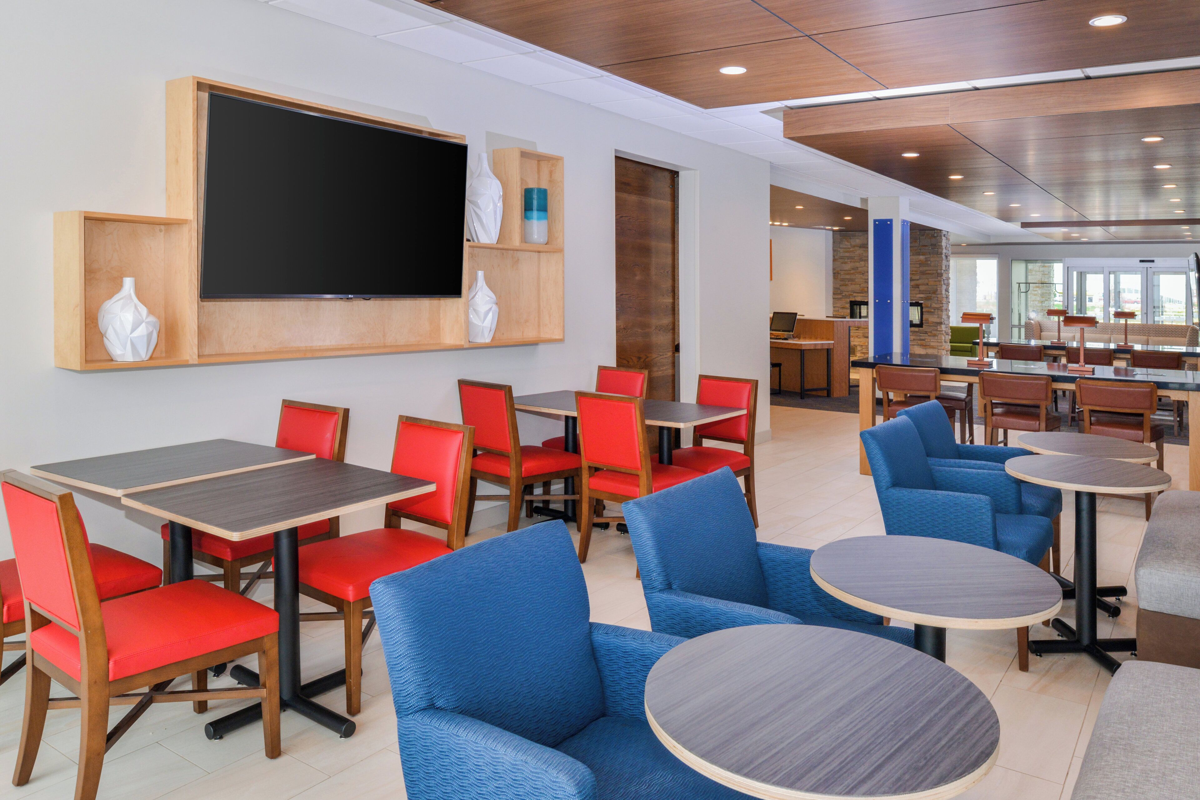 A modern dining area with red and blue seating, a large flat-screen TV, and a kitchen in the background.