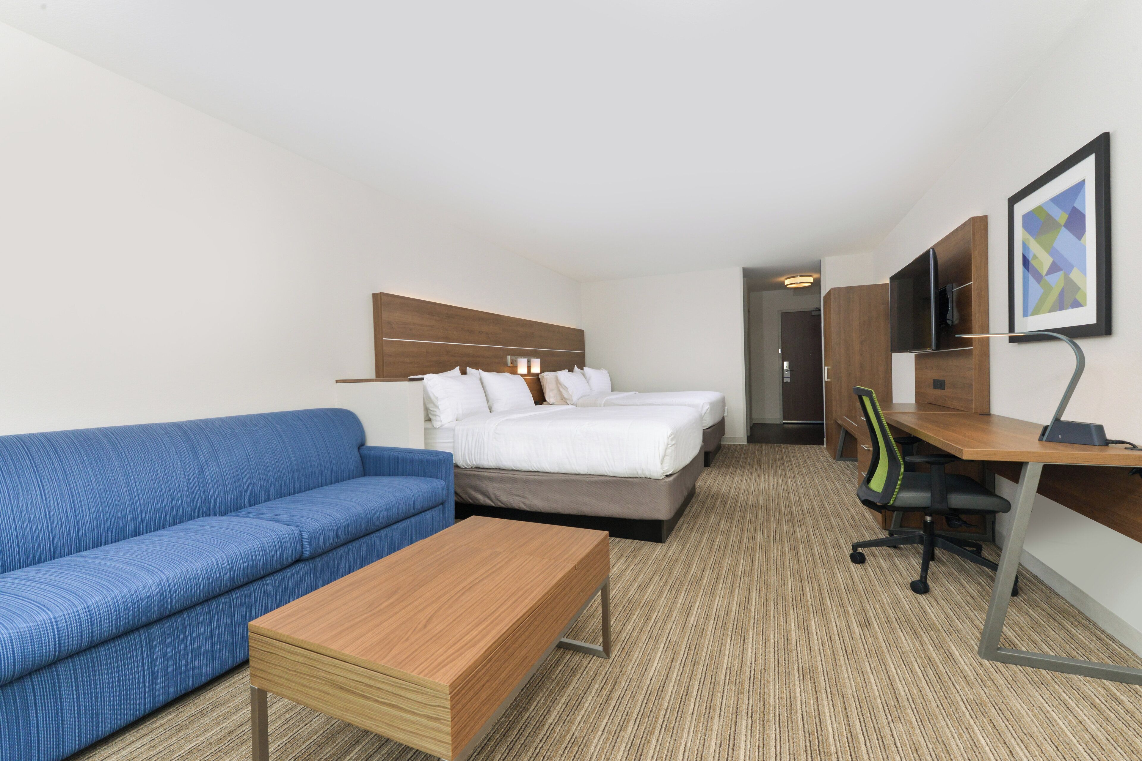 A hotel room with a bed, a blue sofa, a wooden coffee table, a desk with a chair, and a TV on a stand.