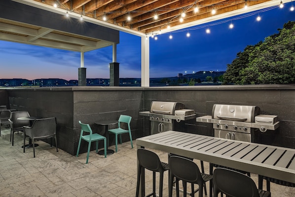 Une terrasse extérieure équipée d’un barbecue, de sièges et offrant une vue sur le paysage urbain au crépuscule.