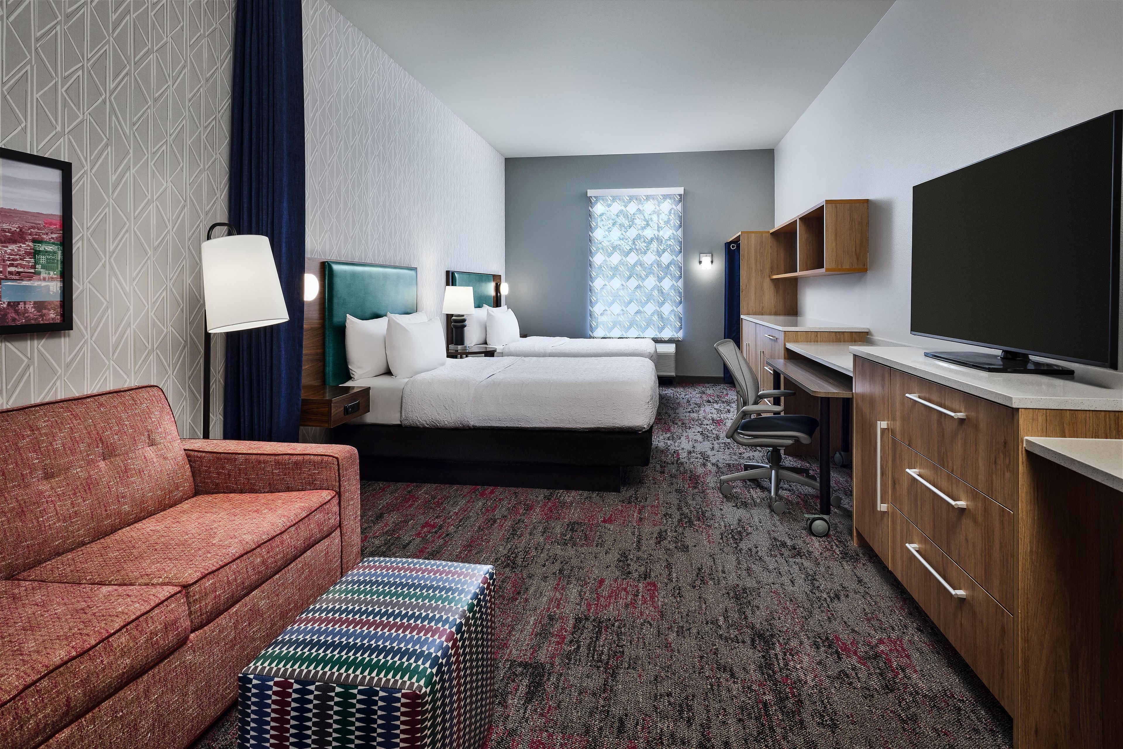 Ein Hotelzimmer mit einer Couch, einem Bett, einem Fernseher, einem Schreibtisch und einem Fenster mit gemusterten Vorhängen.