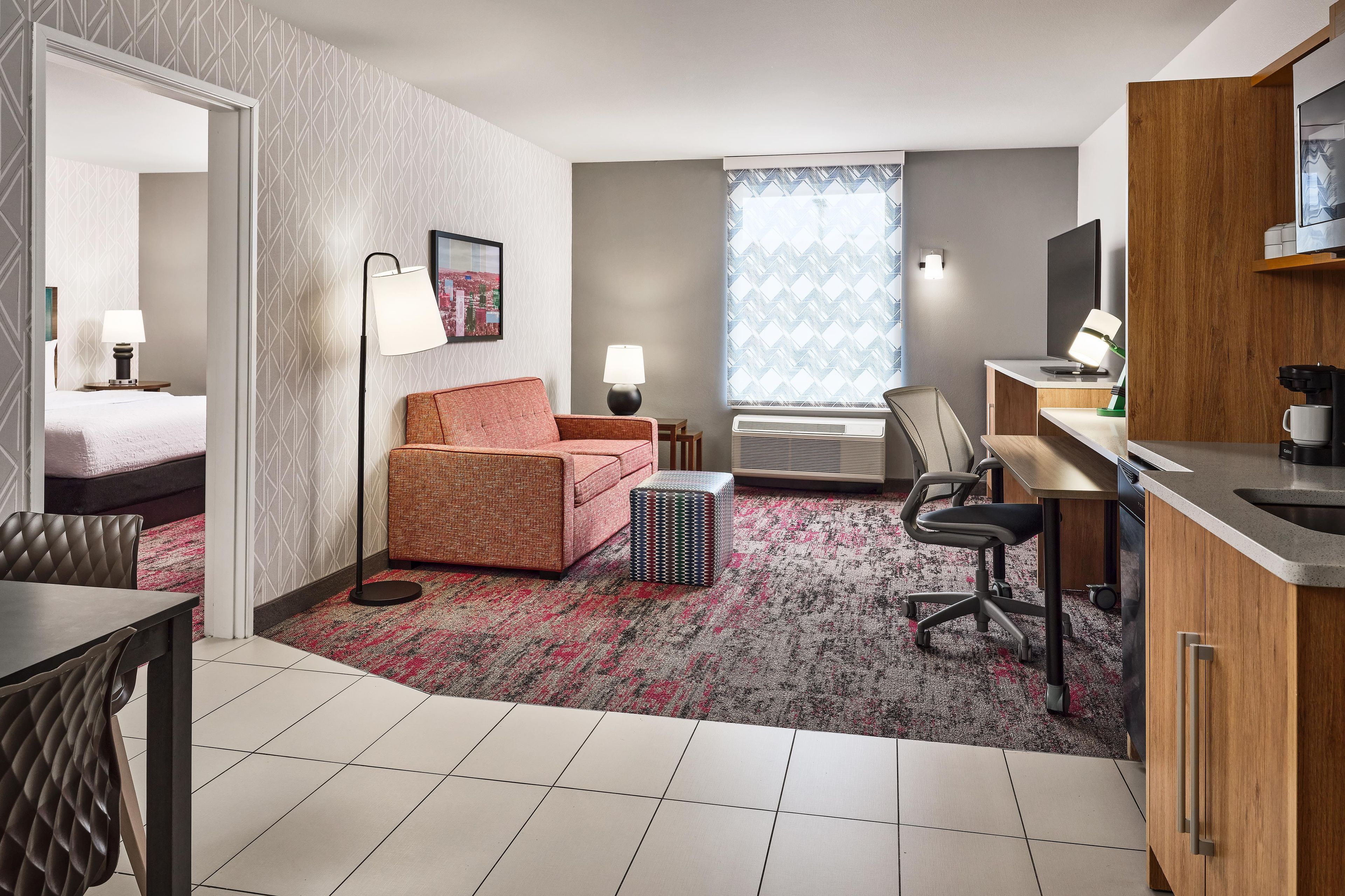 A hotel room with a bed, a sofa, a desk with a computer, a coffee machine, and a window with patterned curtains.
