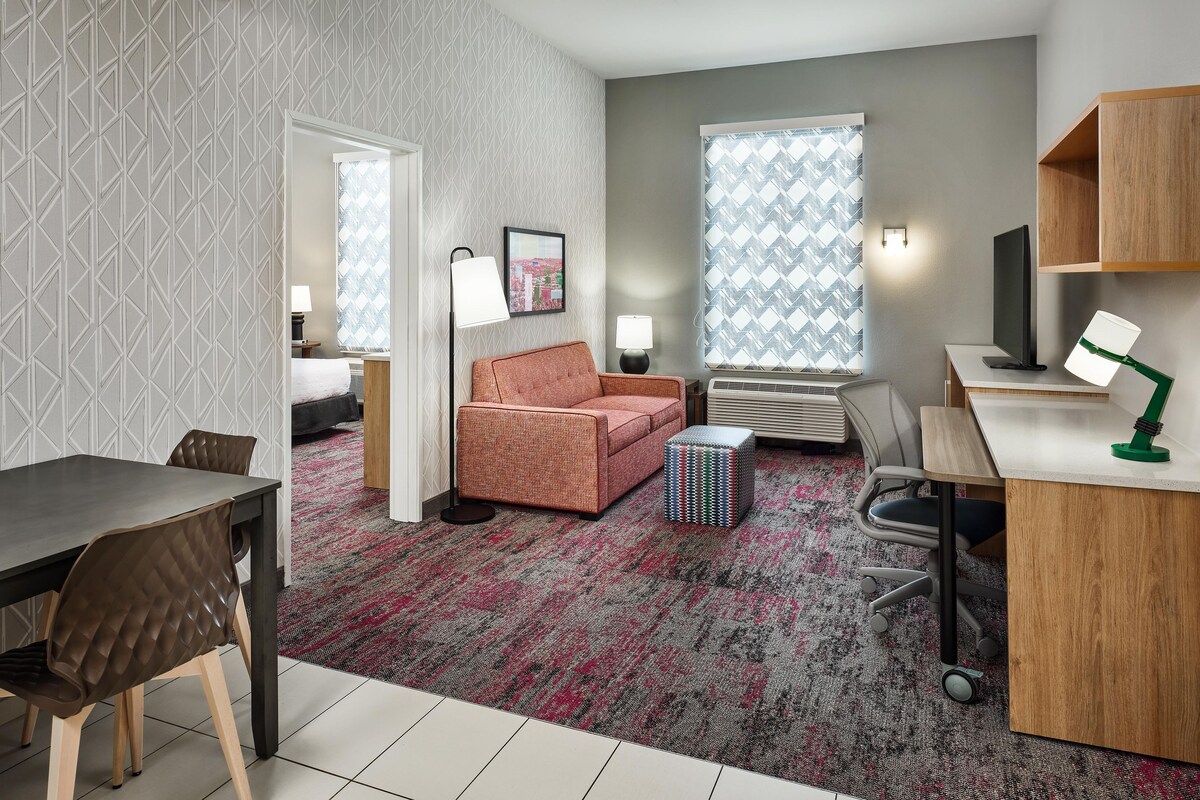 A modern hotel room with a bed, a sofa, a desk, and a chair. There is a window with patterned curtains, a lamp, and a picture on the wall.