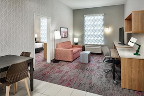 A modern hotel room with a bed, a sofa, a desk, and a chair. There is a window with patterned curtains, a lamp, and a picture on the wall.