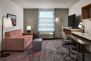 A compact hotel room with a sofa, a desk, a chair, a television, and a patterned window. - Home2 Suites by Hilton Rapid City (Rapid City)
