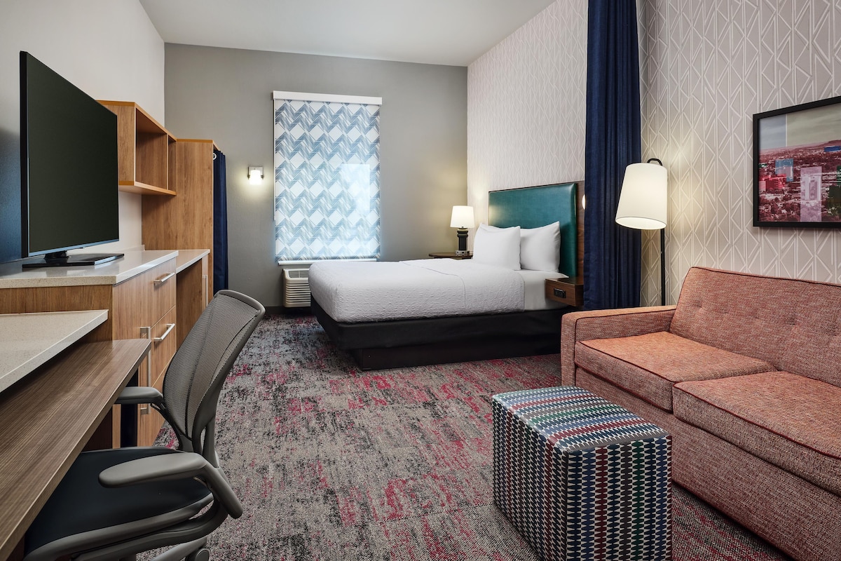 A hotel room with a bed, a desk with a TV, a chair, a sofa, a patterned rug, and a small table.