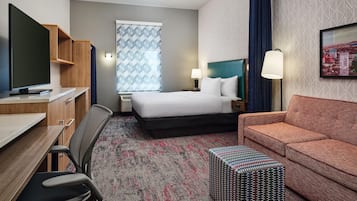 A hotel room with a bed, a desk with a TV, a chair, a sofa, a patterned rug, and a small table.