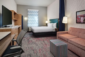 A hotel room with a bed, a desk with a TV, a chair, a sofa, a patterned rug, and a small table.