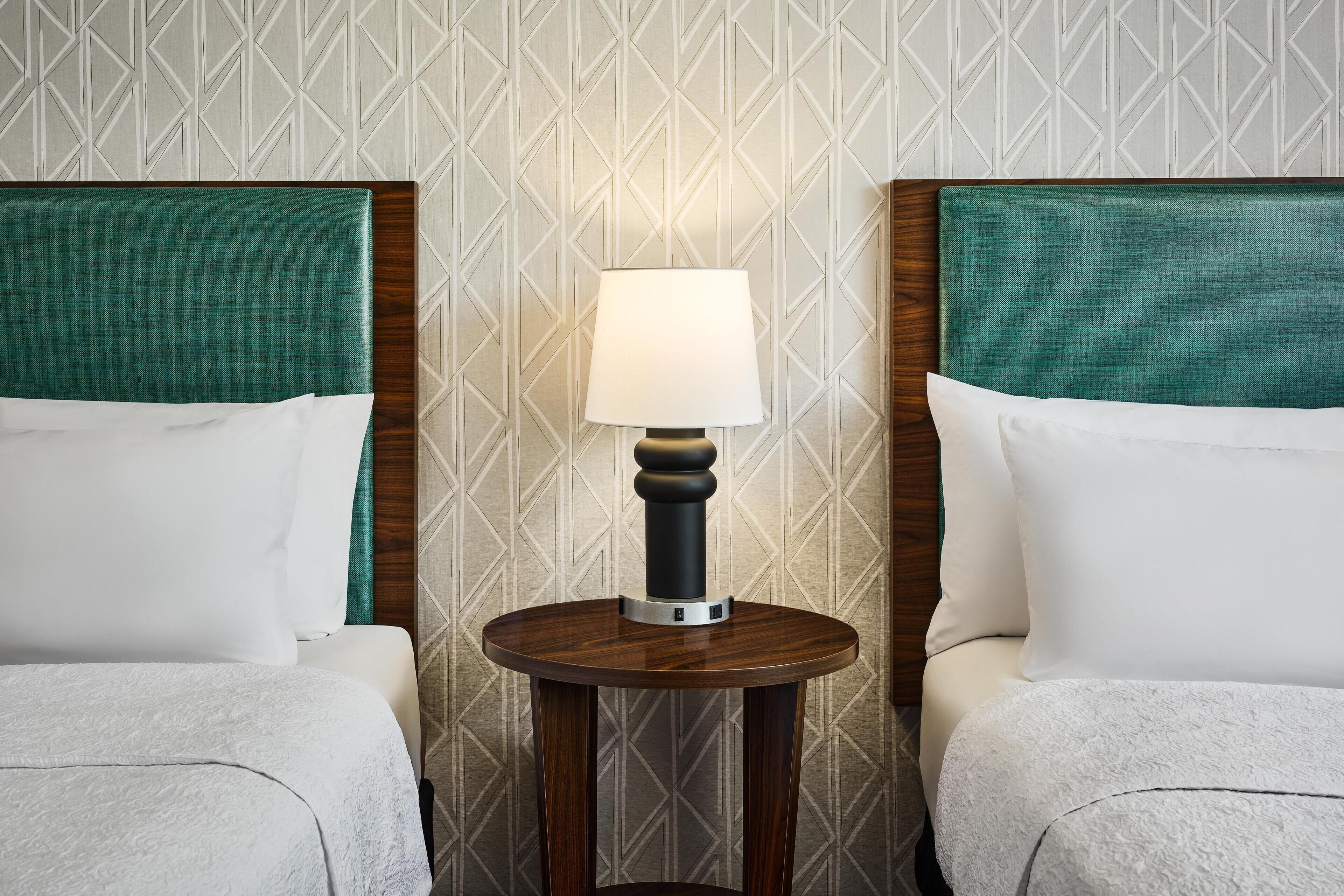 Ein Hotelzimmer mit zwei Betten, einem runden Holztisch und einer Lampe, sowie einer Wand mit Musterung.
