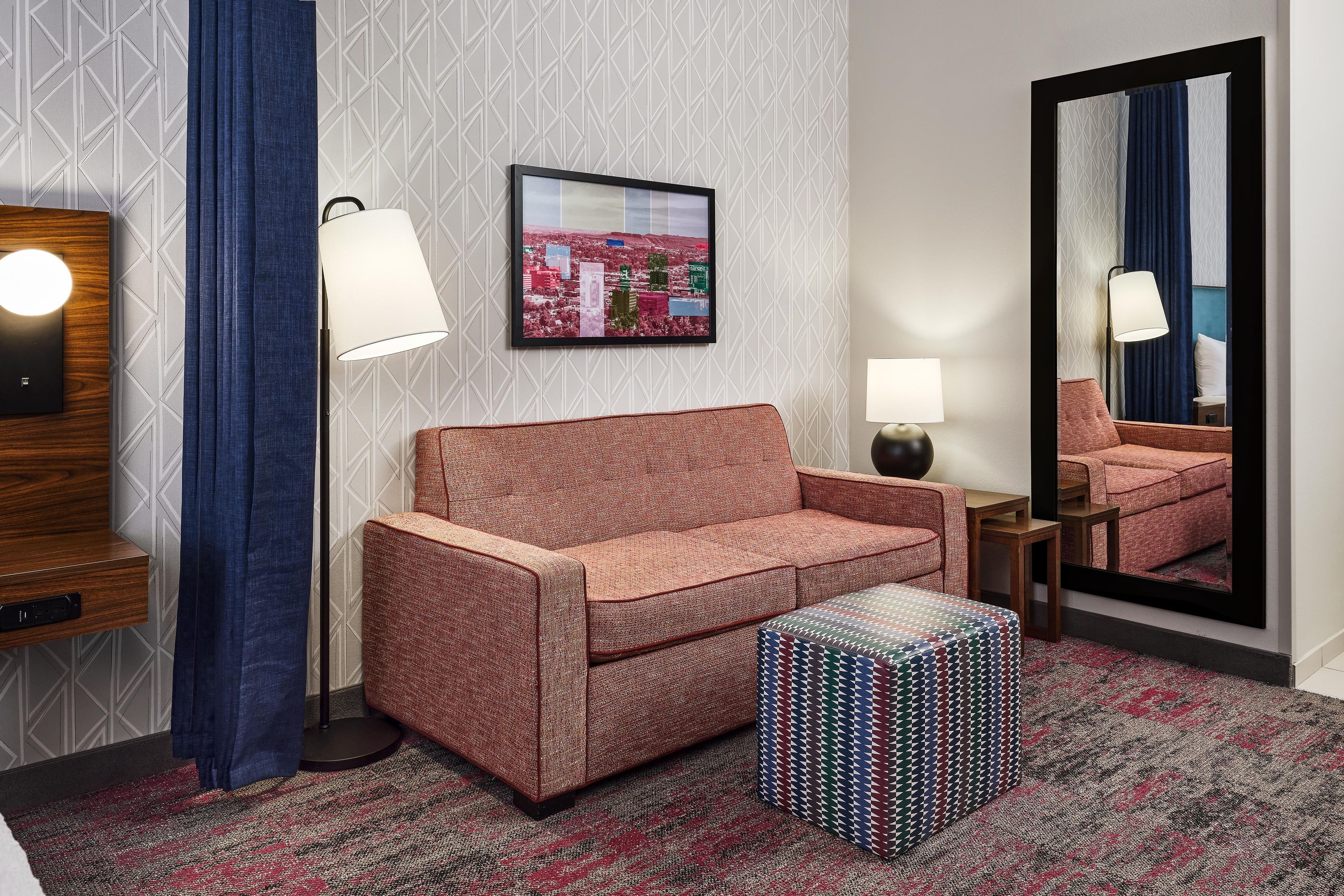 A hotel room with a sofa, a patterned ottoman, a framed cityscape picture, a mirror, and a lamp.