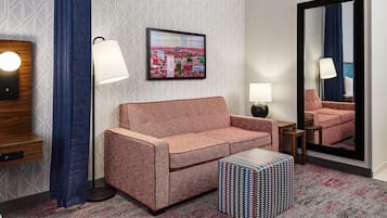 A hotel room with a sofa, a patterned ottoman, a framed cityscape picture, a mirror, and a lamp.