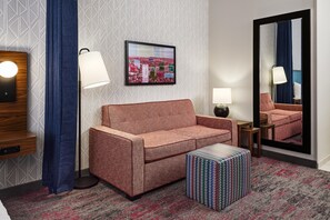A hotel room with a sofa, a patterned ottoman, a framed cityscape picture, a mirror, and a lamp. - Home2 Suites by Hilton Rapid City (Rapid City)