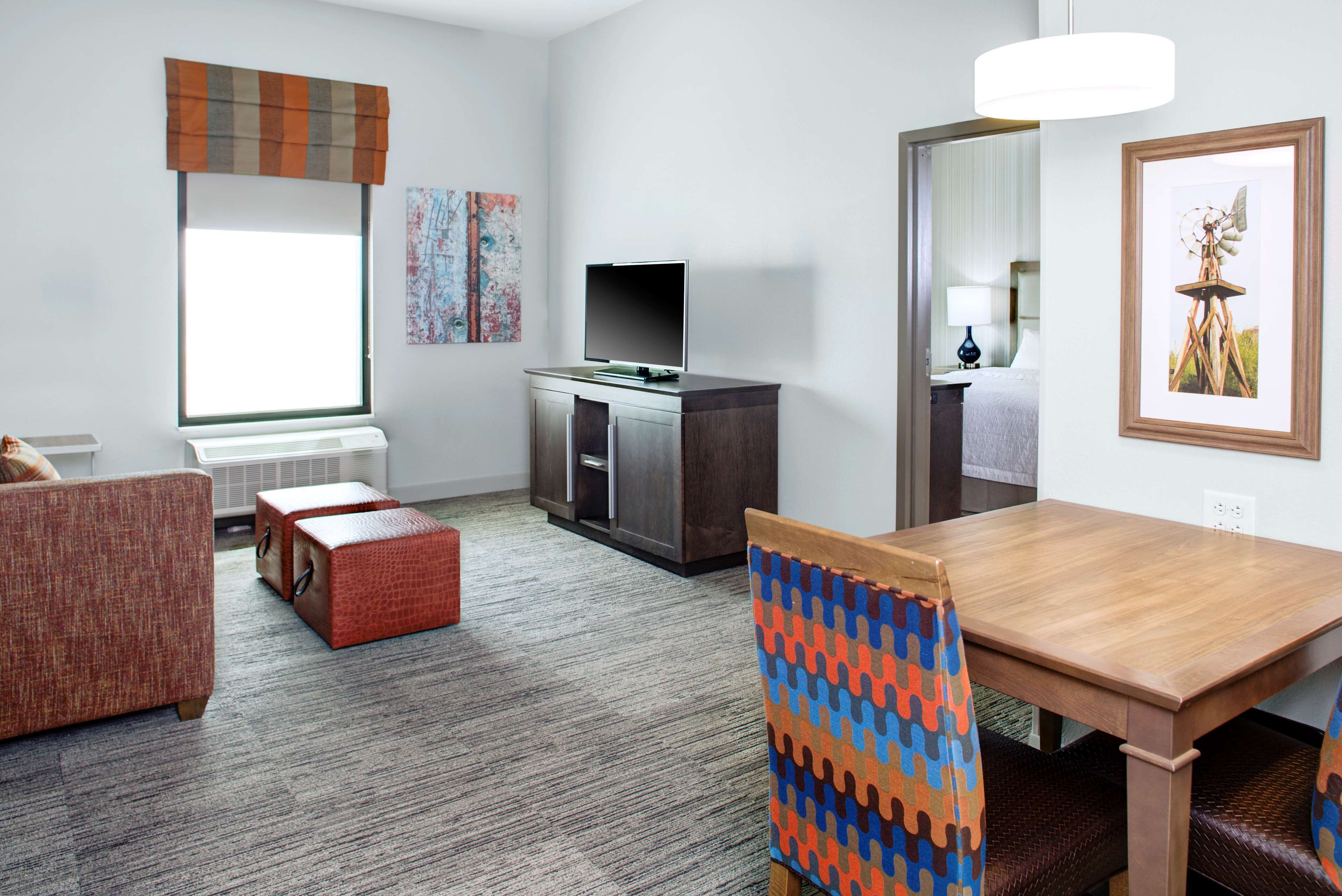 A modern hotel room with a wooden dining table, a TV on a cabinet, a sofa, and a window with blinds.