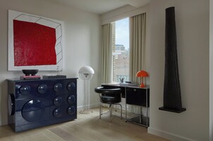 Een moderne, minimalistische kamer met een groot raam, een bureau, een stoel, een hoge zwarte sculptuur en een blauwe kast met een abstract schilderij in rood.