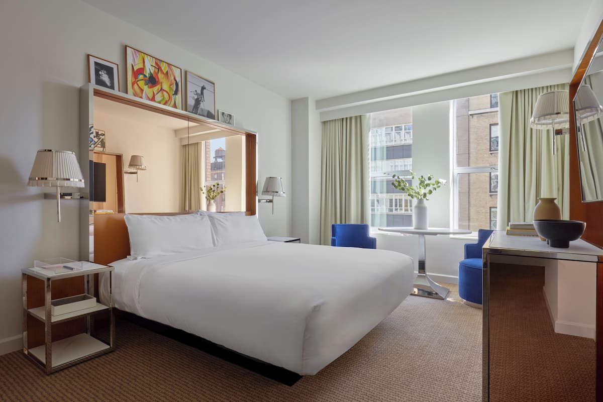 Habitación de hotel con una cama grande, un escritorio con lámpara, una silla, una mesita con un florero y una ventana con cortinas.