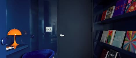 A room with a dark blue door, a blue chair, a small orange table lamp, and a bookshelf with various books.