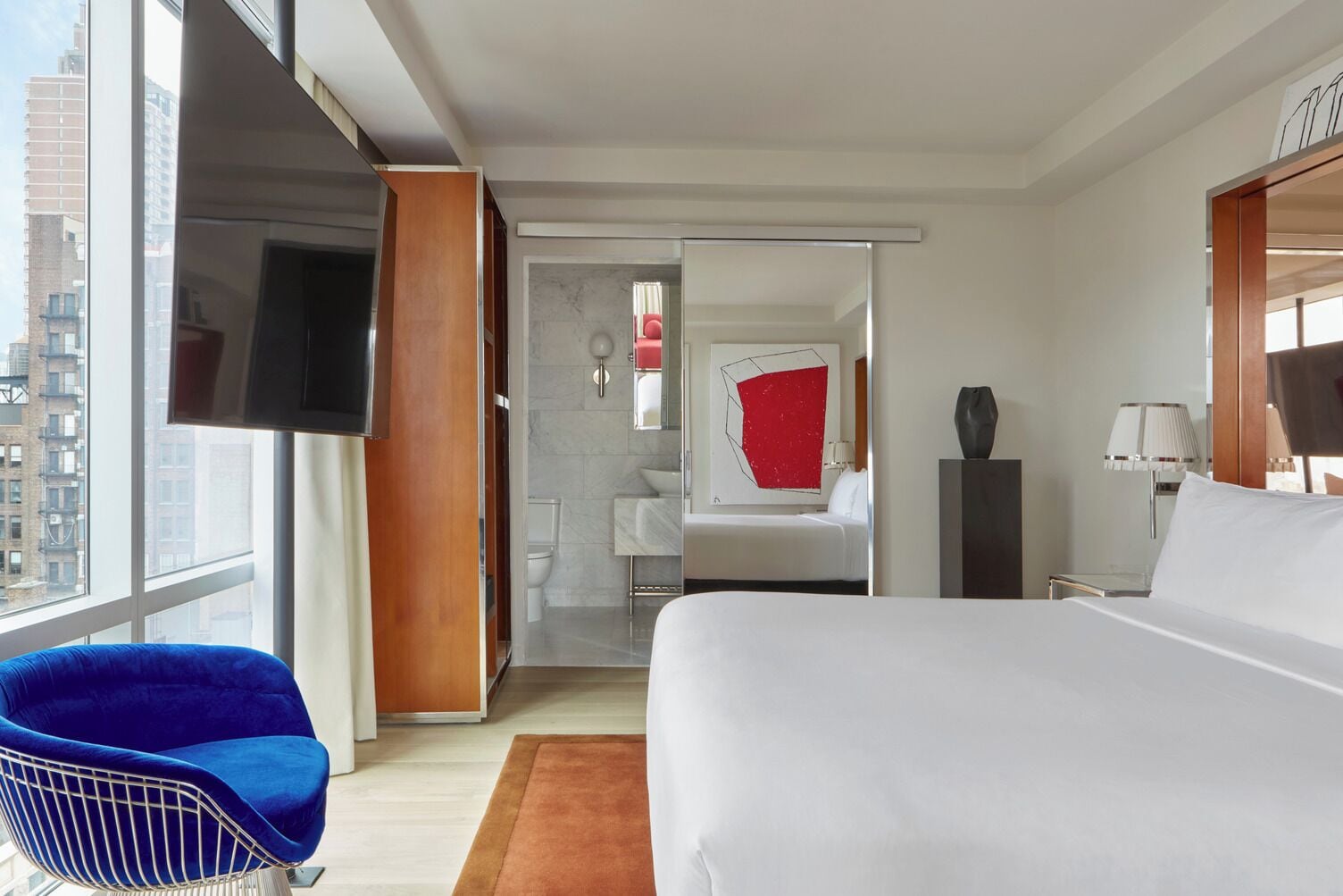 A modern hotel room with a large bed, a blue chair, a bathroom with a red accent wall, and a city view.