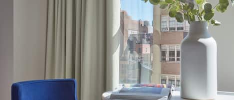 A modern room with a blue armchair, a white table with a vase of green plants, and a view of buildings through a window with beige curtains.