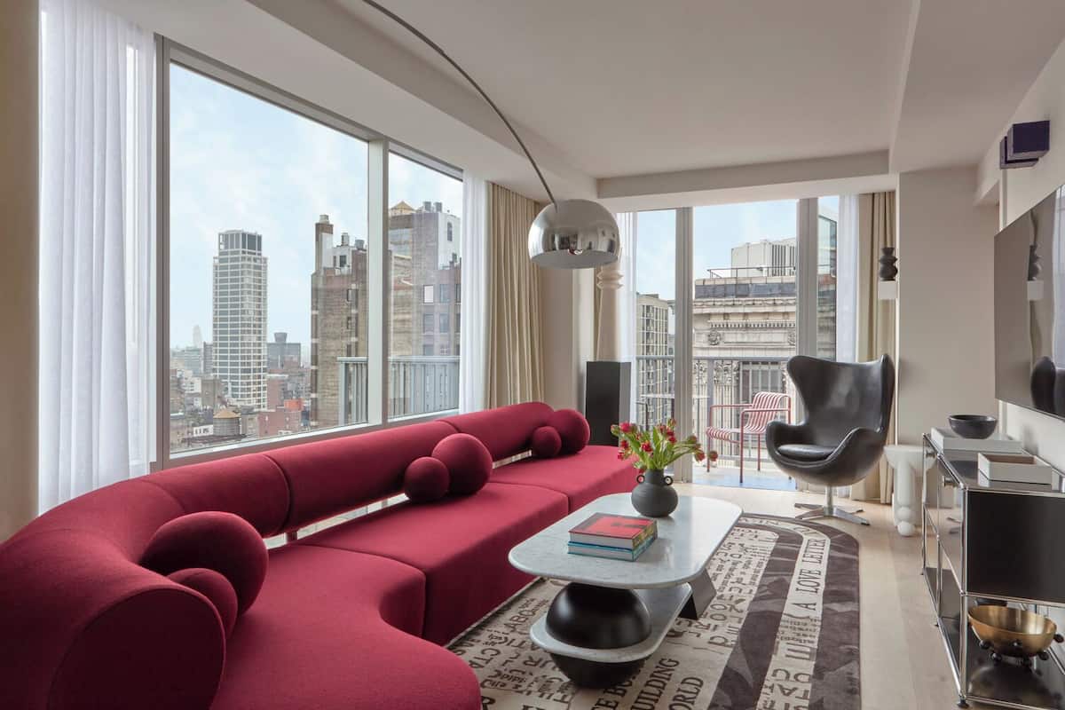 Una sala de estar moderna con un sofá sectional rojo, un sillón negro, una mesa de centro con libros y vistas de la ciudad a través de ventanas grandes.