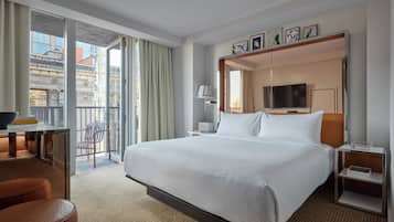 Habitación de hotel con una cama grande, un escritorio con silla, televisor y balcón con vistas a edificios.