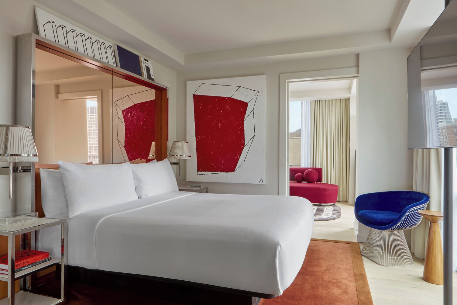 A modern hotel room with a large bed, a red accent wall, a red chair, and a blue chair.