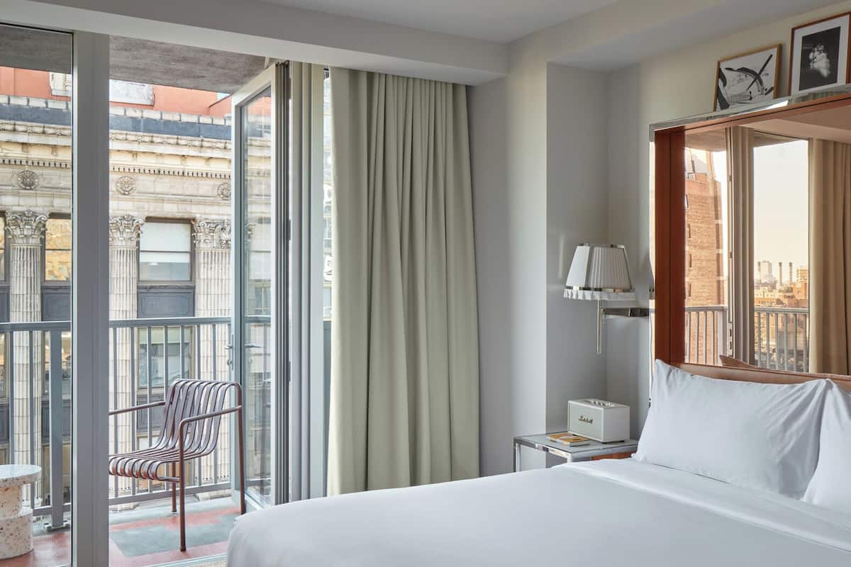 Habitación de hotel con una cama grande, una silla, una mesita de noche, una lámpara y vistas a la ciudad desde la ventana.