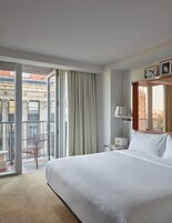 Habitación de hotel con una cama grande, una silla, una mesita de noche, una lámpara y vistas a la ciudad desde la ventana.