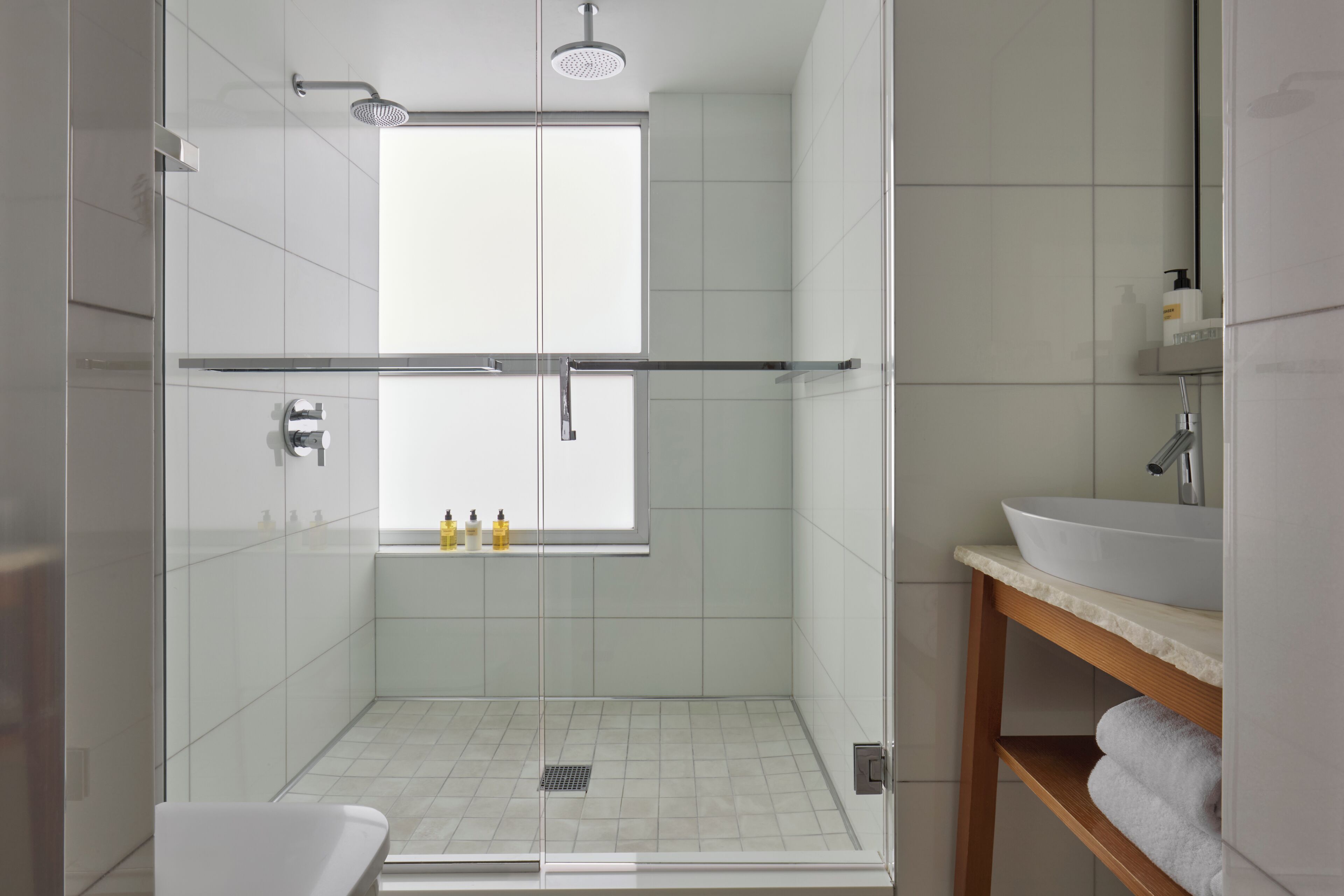 Un baño moderno con ducha de vidrio, un lavamanos blanco y una ventana sobre la ducha.