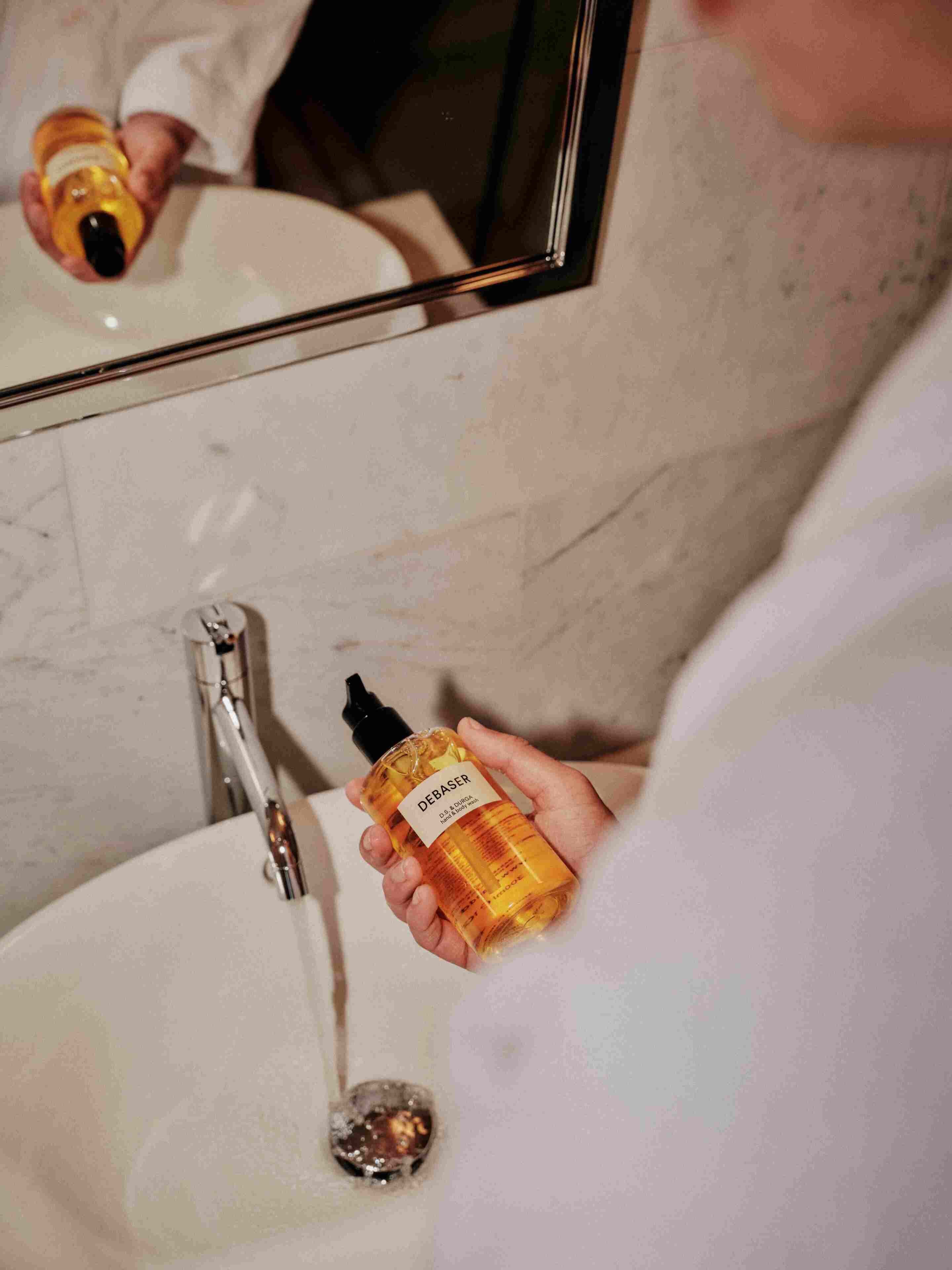 A person holding a bottle of DEBASER next to a bathroom sink with a mirror above it.