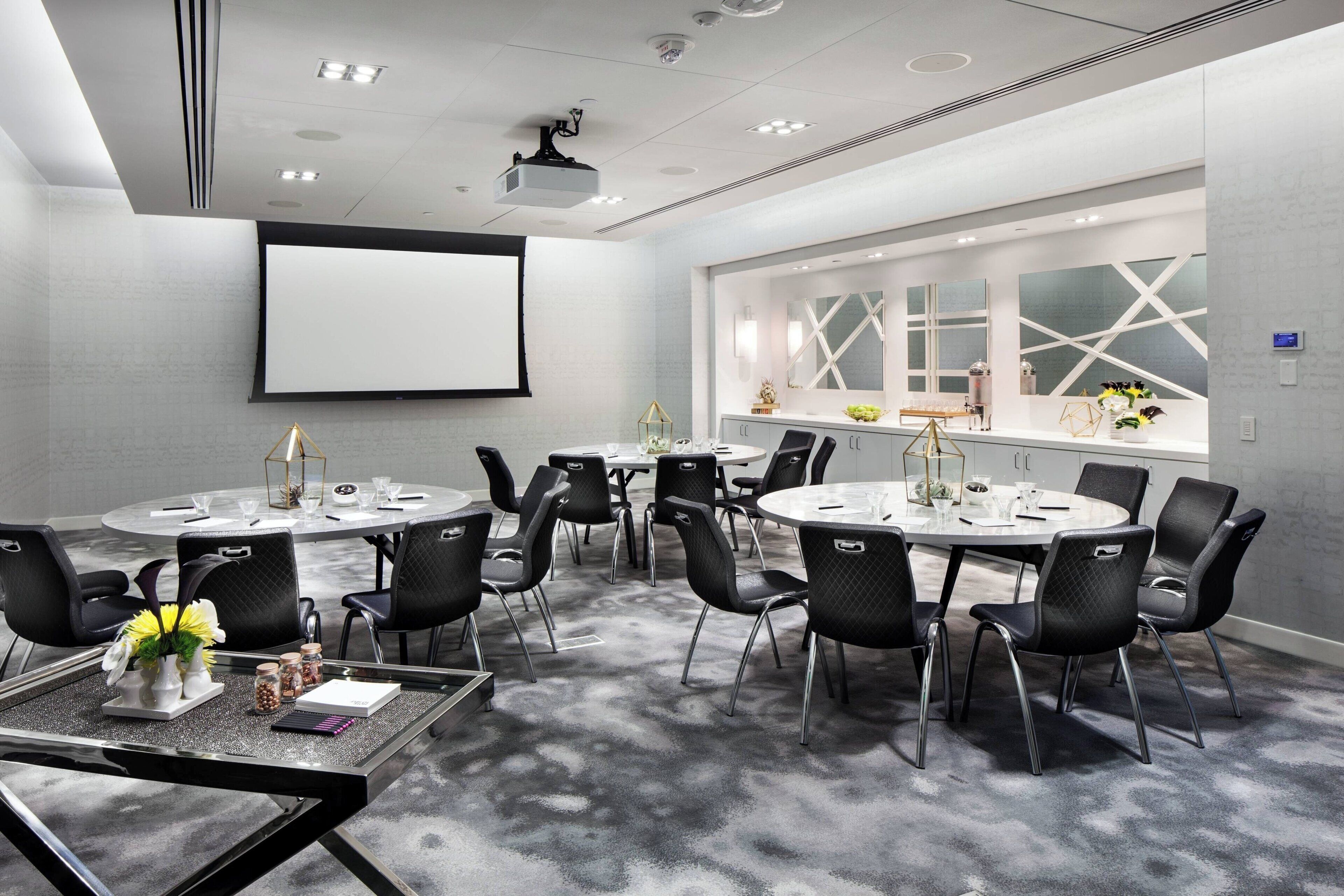 A conference room with round tables, black chairs, a projector screen, and a large window with geometric patterns.