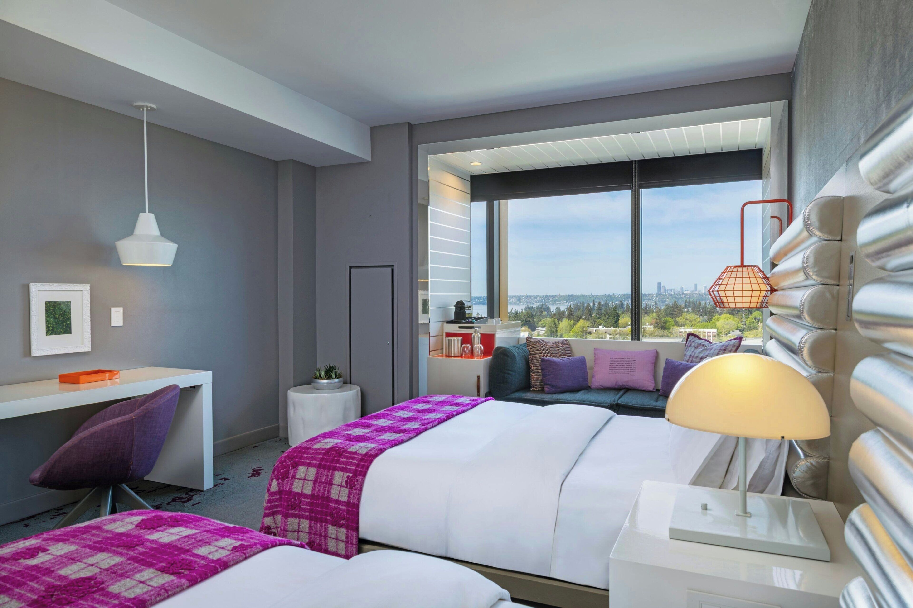 Habitación de hotel con dos camas, un escritorio, una silla, una lámpara y un ventanal con vistas a la ciudad.