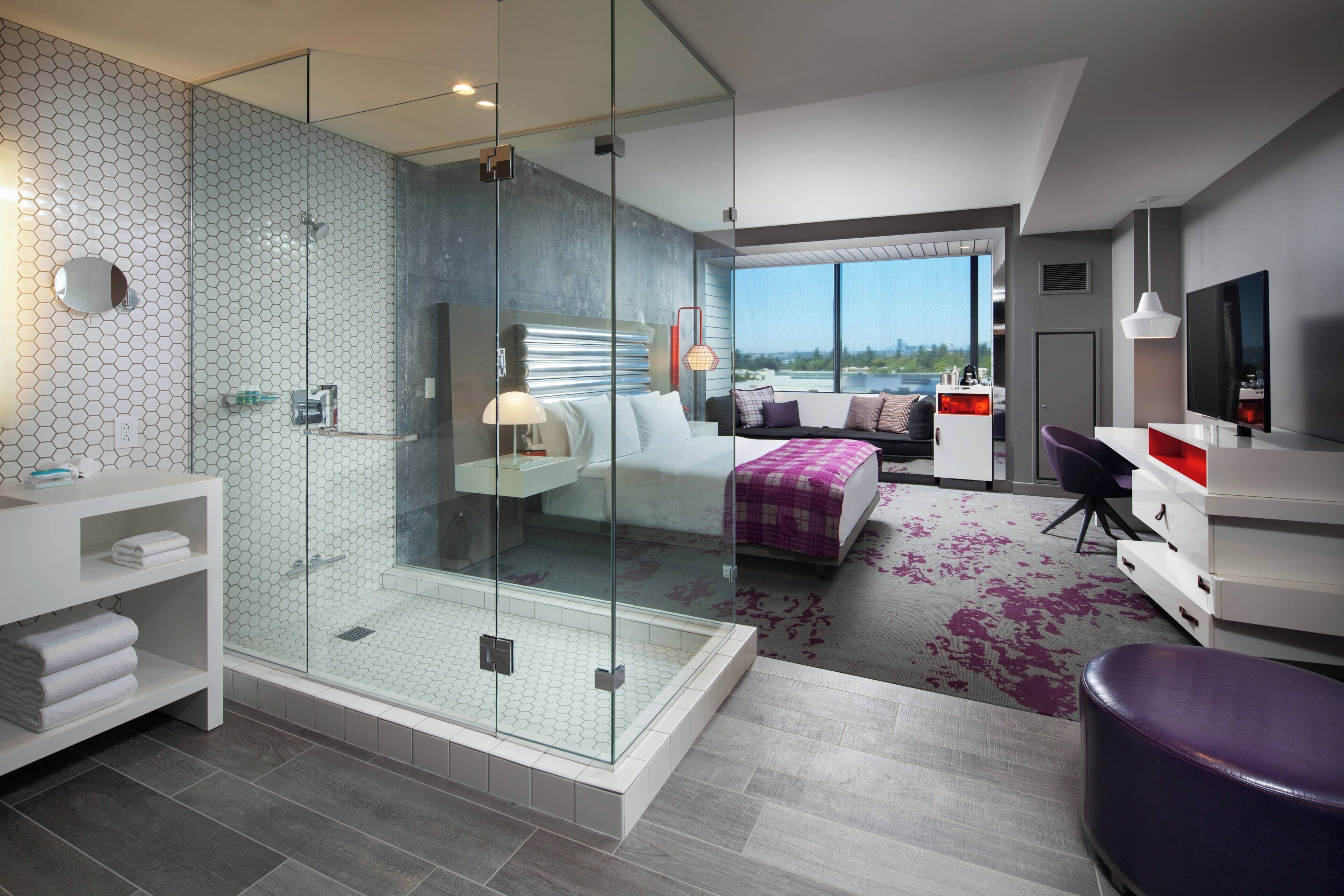 Ein modernes Hotelzimmer mit einer gläsernen Duschkabine, einem Bett mit Blick, einem Schreibtisch mit Stuhl und einem großen Fenster.