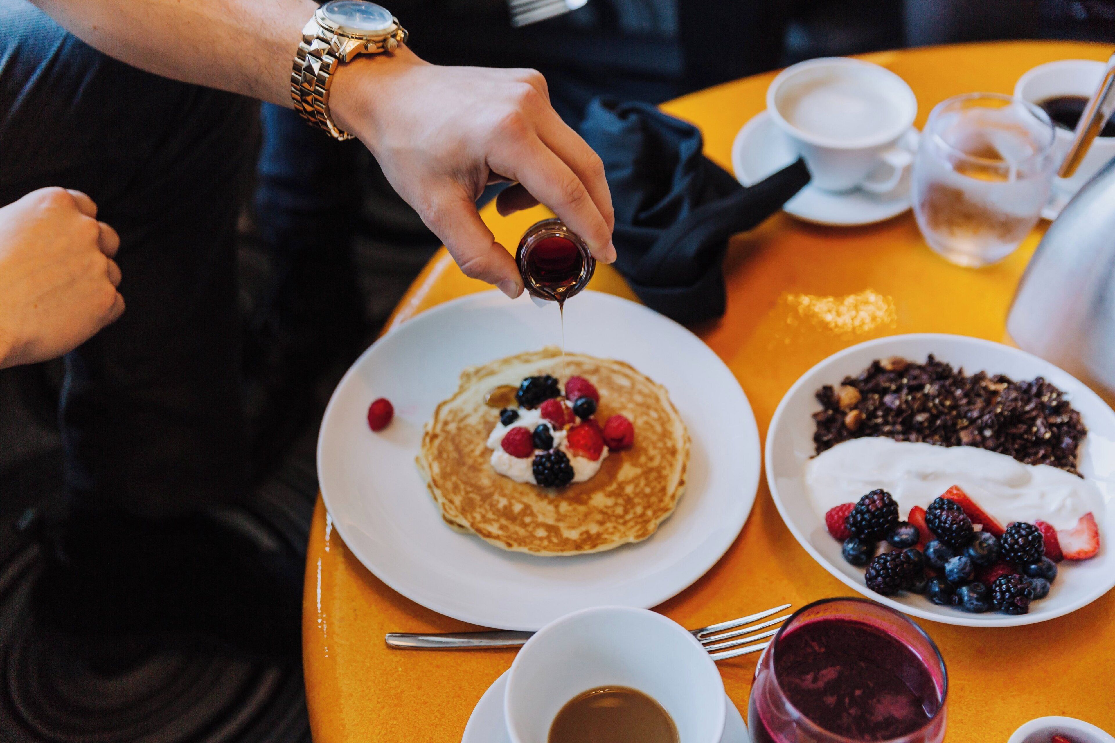 A person pouring syrup onto pancakes with berries on a table with coffee and a bowl of yogurt with berries.