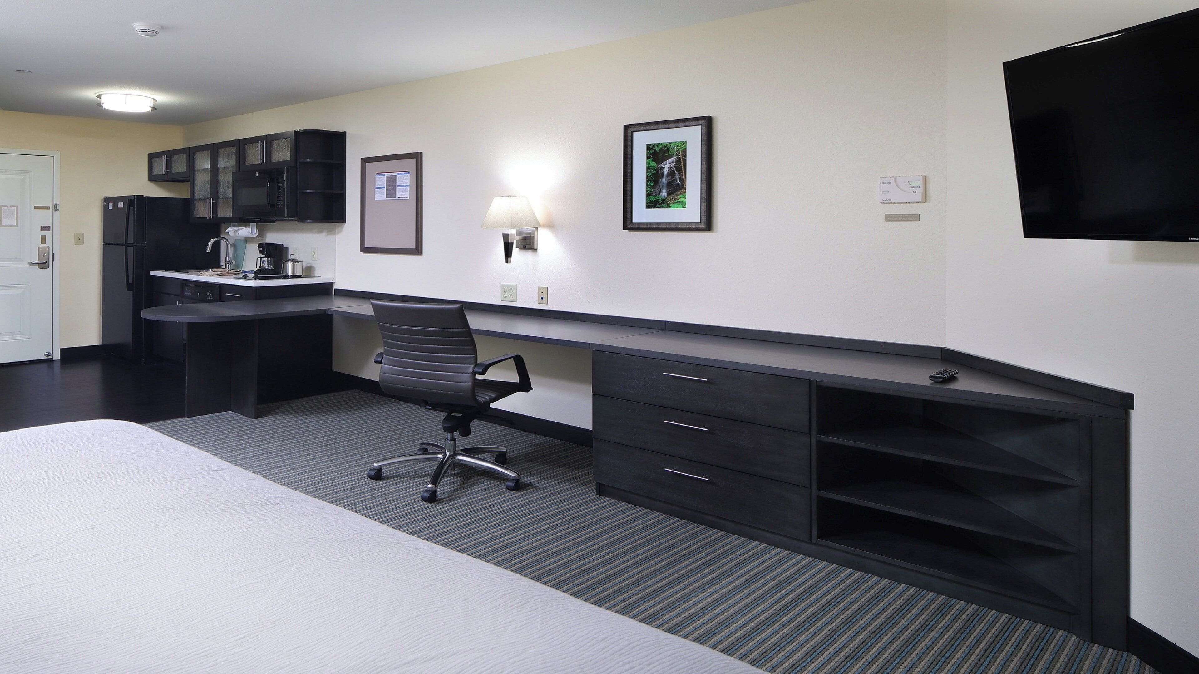 A hotel room with a bed, desk, chair, kitchenette, and a TV.