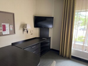 A hotel room with a flat-screen TV mounted on the wall, a desk with drawers, a lamp, and a bulletin board with safety information.