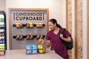 Una persona con uniforme morado está de pie junto a una barra con un cartel que dice 'CANDLEWOOD CUPBOARD' sobre dispensadores de snacks.