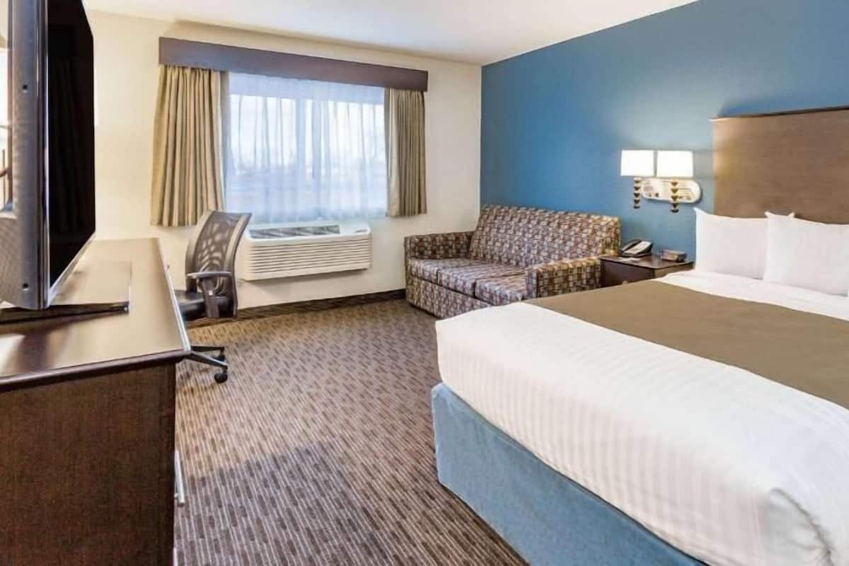 A hotel room with a large bed, a desk with a TV, a chair, a sofa, an air conditioning unit, and a lamp.