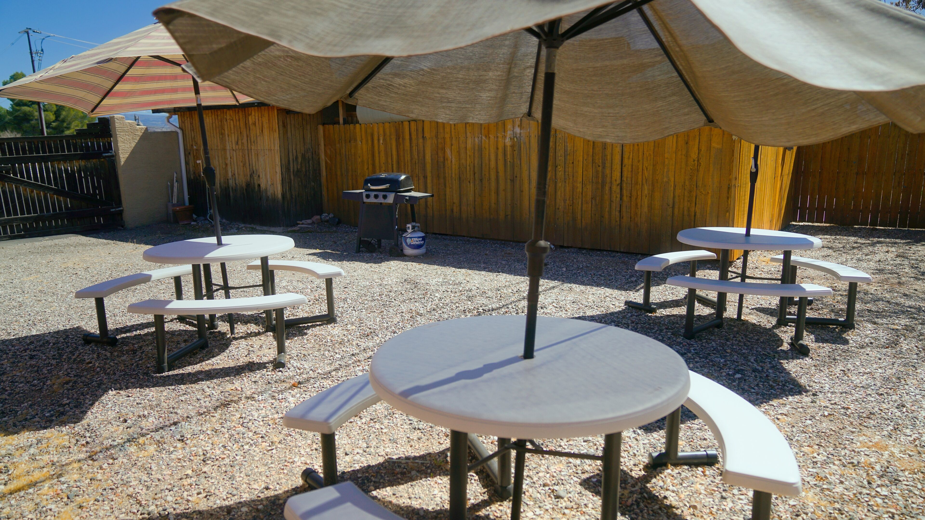 An outdoor patio area with round tables and benches, covered by large umbrellas, and a wooden fence in the background.