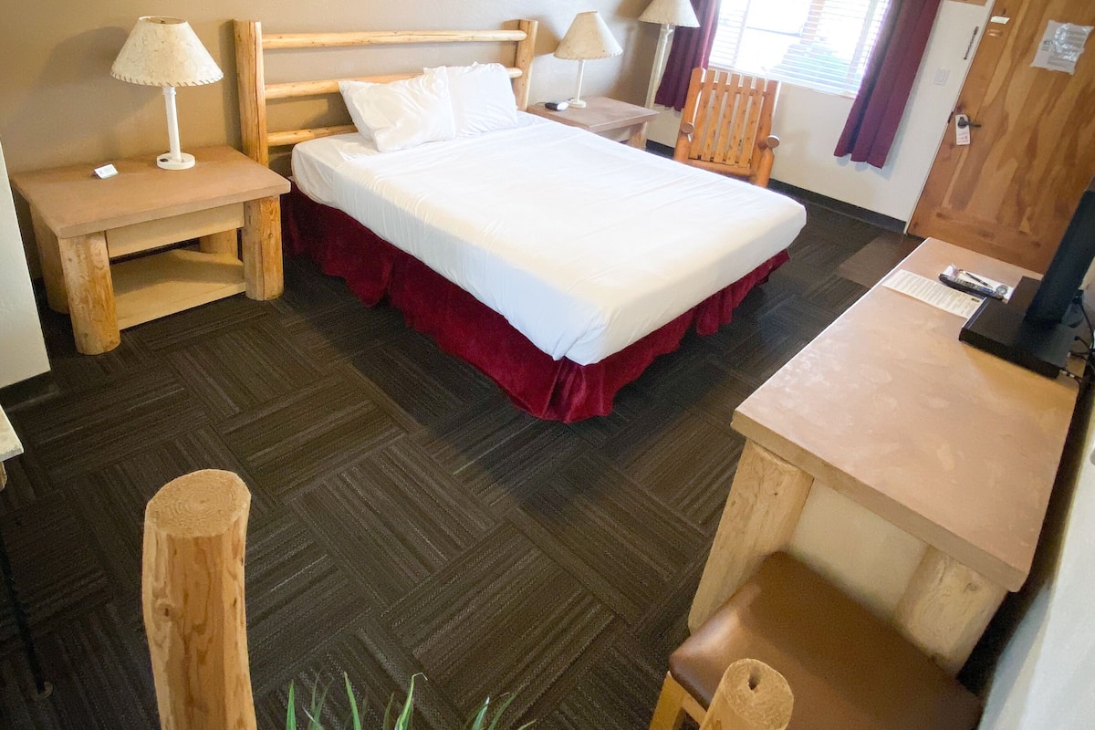 A hotel room with a bed, bedside tables, a desk, a chair, a ceiling fan, and a window with curtains.