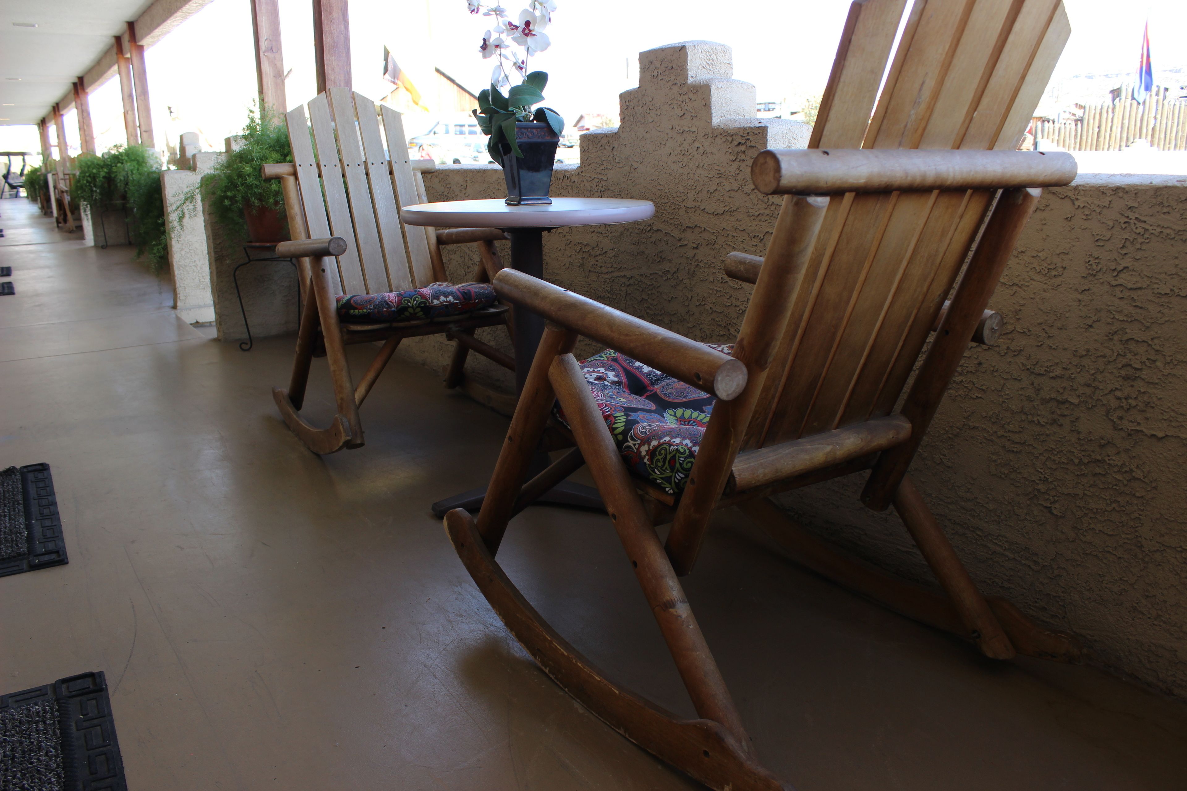 A covered outdoor seating area with wooden rocking chairs and a small table, surrounded by potted plants and a textured wall.