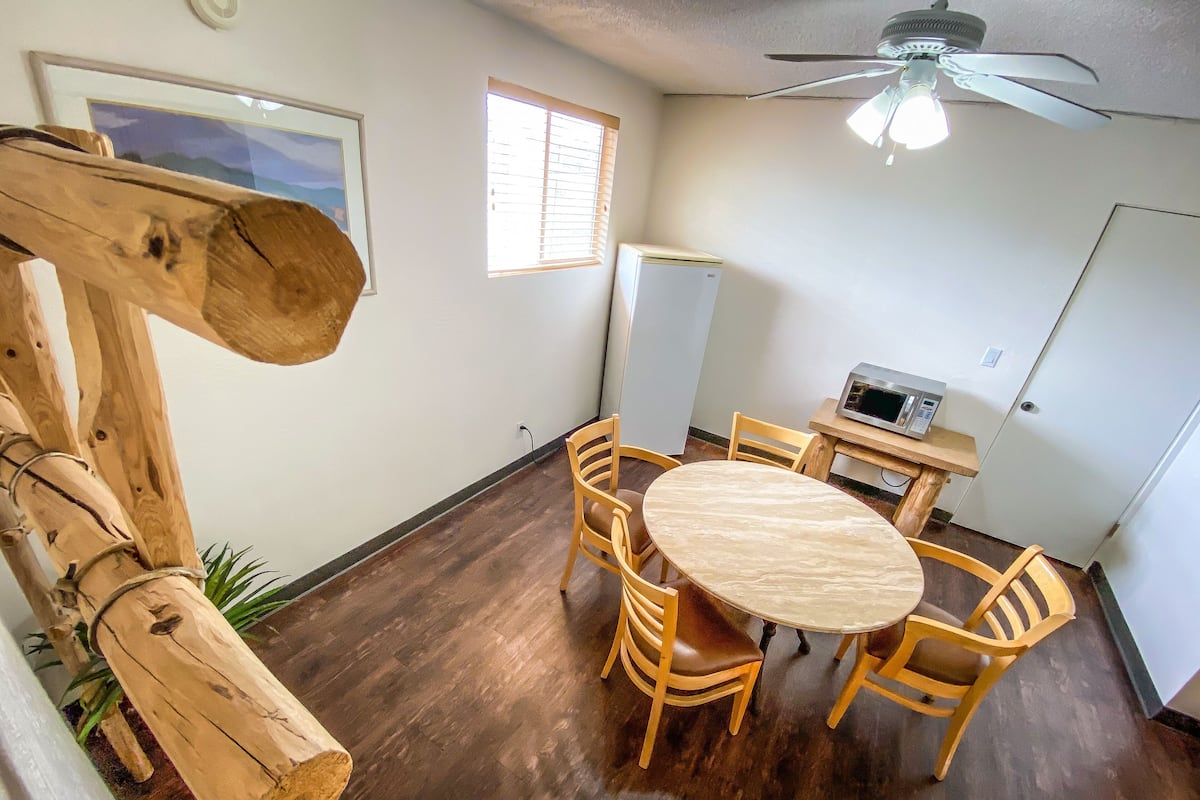 A dining area with a round table and wooden chairs, a ceiling fan, a window with blinds, and a refrigerator.