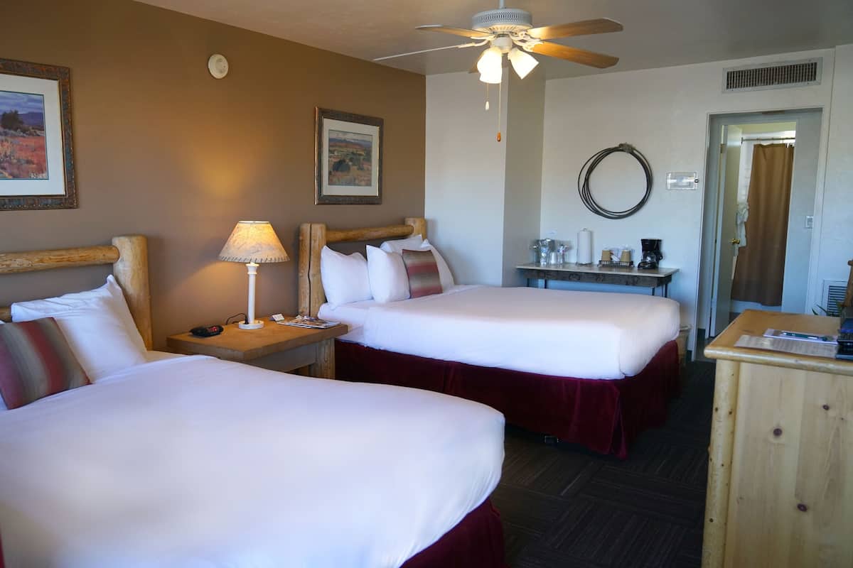 A hotel room with two beds, a desk, a ceiling fan, and two framed pictures on the wall.