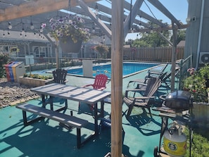 Un espace aménagé au bord de la piscine, avec une table et des chaises, un pot de fleurs suspendu et une bouteille de propane.