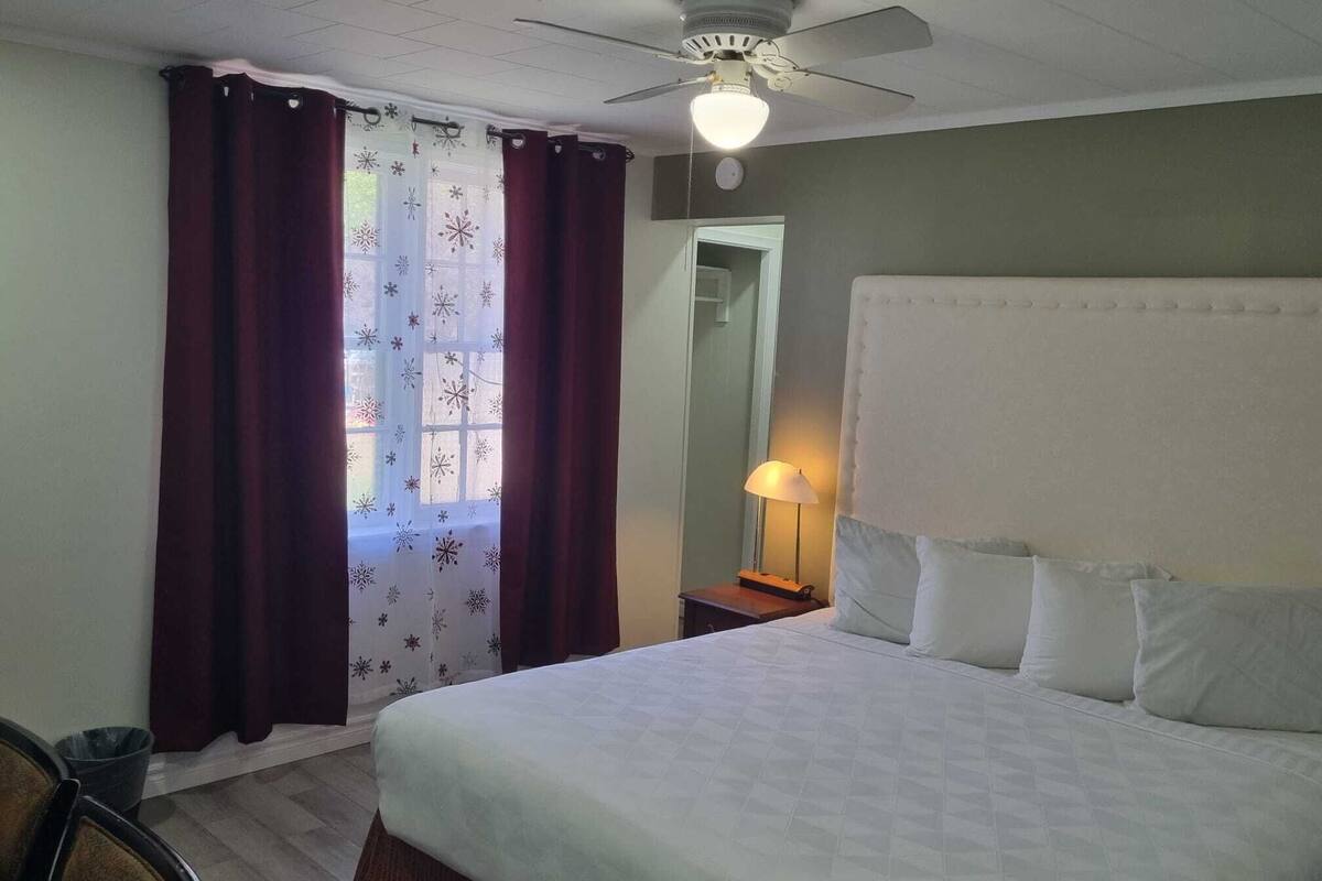 Quarto de hotel com cama, mesa de cabeceira, luminária, ventilador de teto e janela com cortinas.