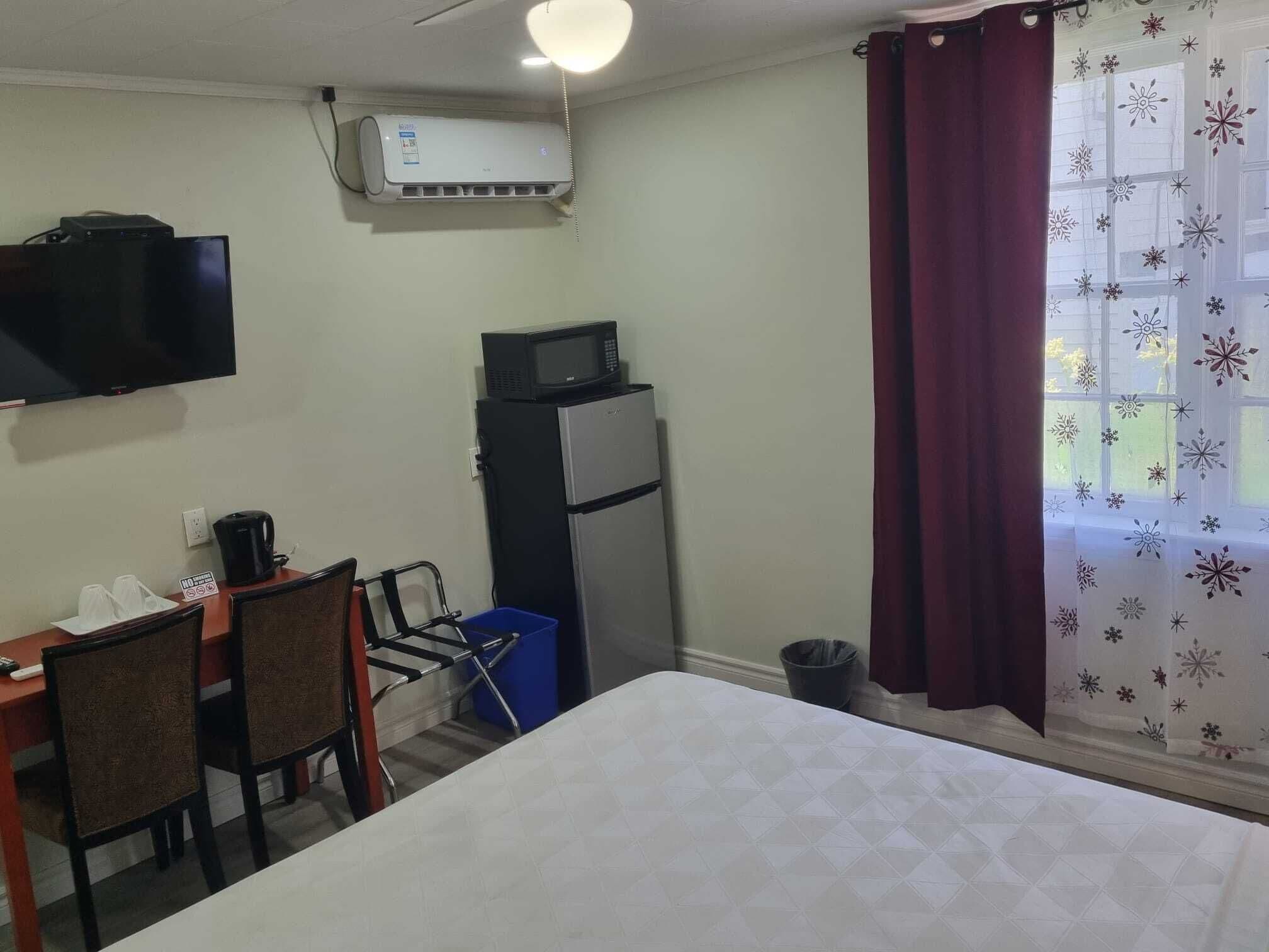 Una habitación con cama, escritorio y sillas, televisor, microondas, refrigerador y ventana con cortinas.