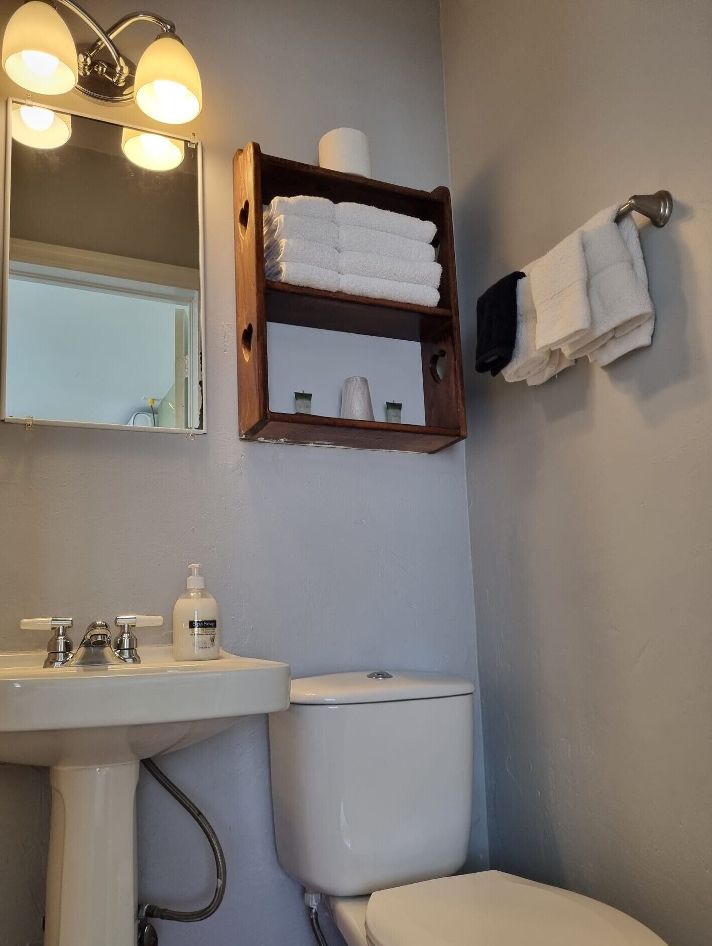 Ein Badezimmer mit Waschbecken, Toilette und einem Holzregal mit Handtüchern und einer Toilettenpapierrolle.