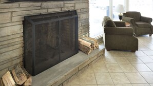 Une cheminée en pierre avec un paravent en métal, une pile de bois de chauffage et deux fauteuils dans une pièce dont le sol est recouvert de carreaux.