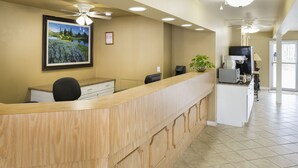Un espace d’accueil avec un comptoir en bois, un ventilateur de plafond, un tableau représentant un paysage encadré, et, au fond, un coin cuisine.