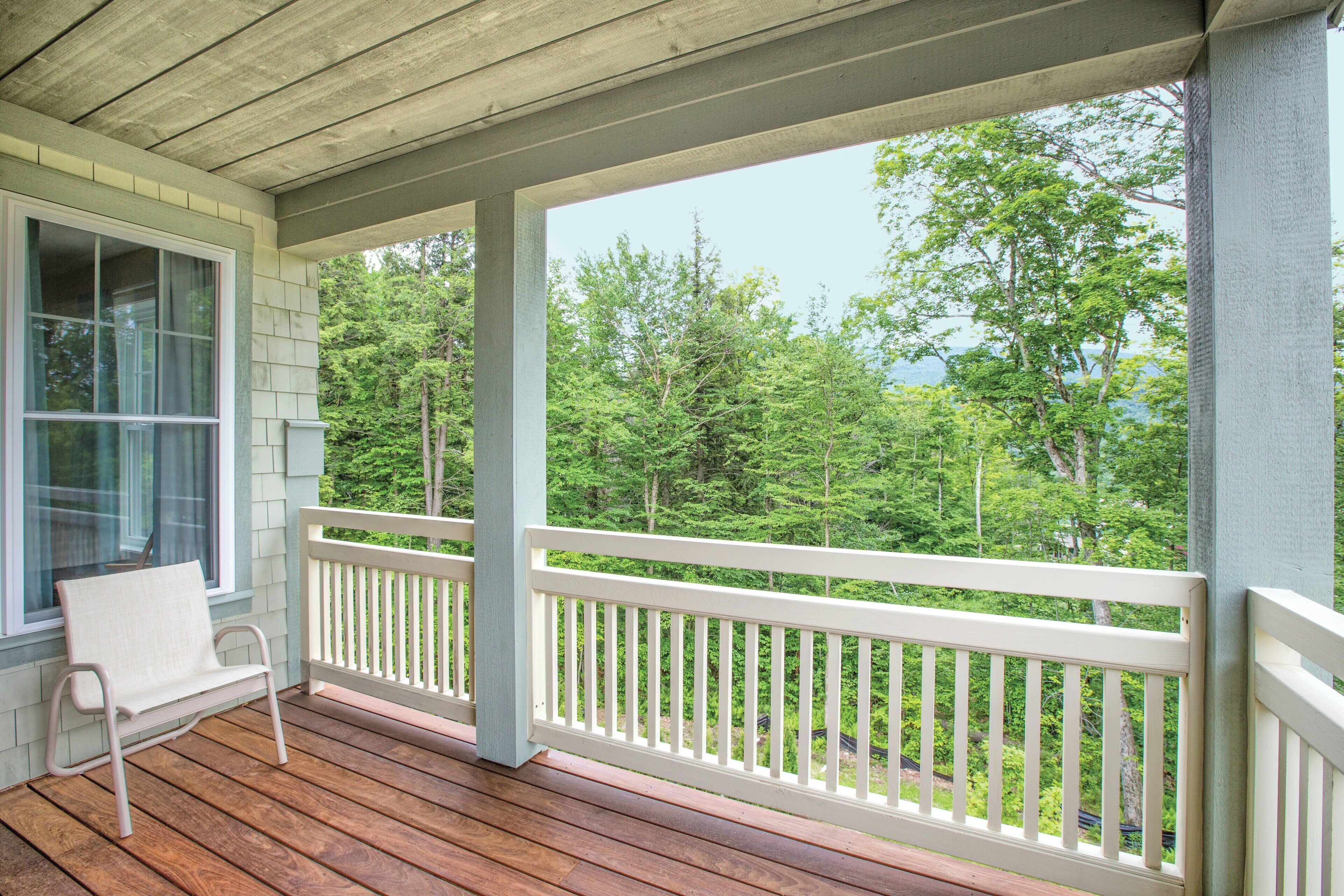 A covered porch with a white railing, a wooden deck, and a view of a forested area.
