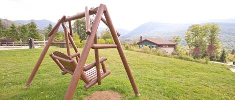 A wooden swing set in a grassy field with a house and mountains in the background.