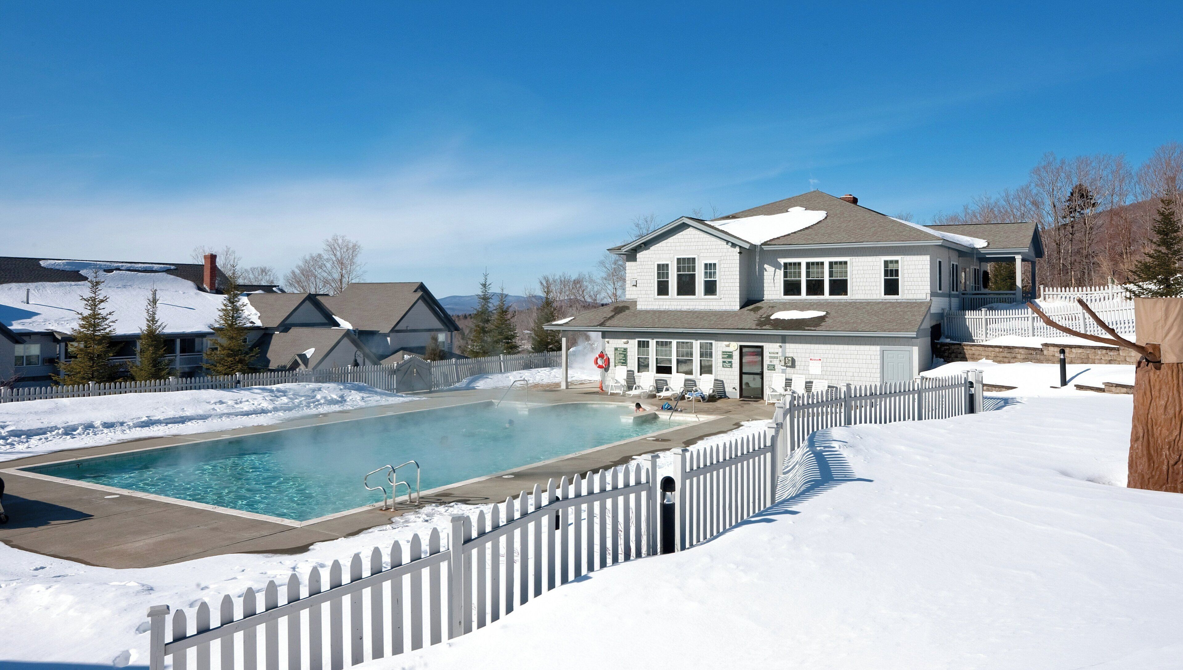 Un quartier résidentiel avec une piscine entourée d’une clôture blanche en bois, une maison dotée d’un porche et un paysage enneigé.